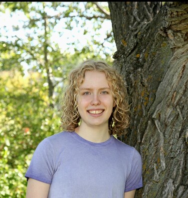 A high school student with shoulder-length blonde hair smiles at the camera while wearing a light purple shirt. To the right there's the trunk of a tree and to the left there's many green leaves from other trees in the background.