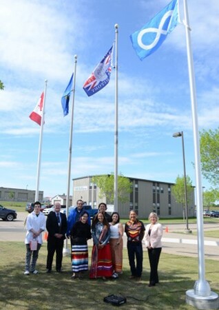 Group of nine adults stand outside in front of four flag poles displaying the Canada, Alberta, Treaty 6 and Métis Nation flags