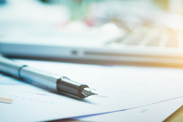 Closeup of a silver ballpoint pen on sheets of paper with a laptop in a blurred background.