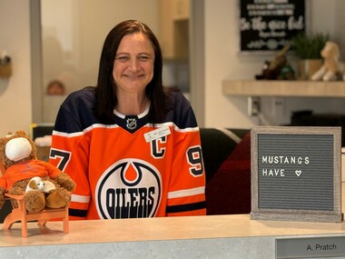 A brown-haired woman sits at a school front office desk and smiles at the camera.