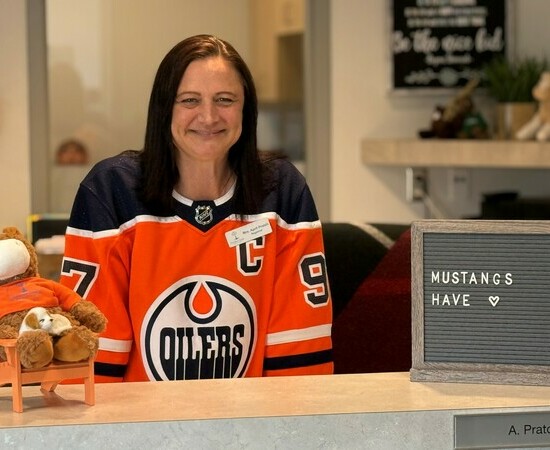 A brown-haired woman sits at a school front office desk and smiles at the camera.