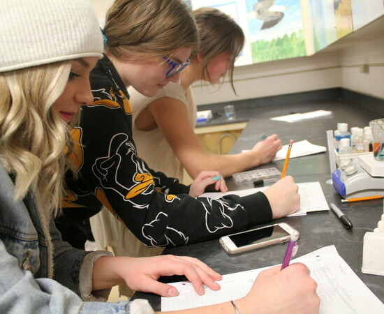 Three students in a science classroom stand at a counter littered with test tubes filled with solutions. There are also notebooks and other school supplies on the counter.