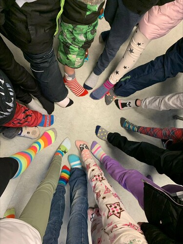 Young students stand in circle on a white floor with their right legs outstretched, sporting socks of various colours, patterns and designs.