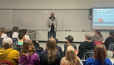 Amit Mali stands in front of a white board with students sitting in chairs in front of him listening. There's a projector screen to the right with Mali's presentation open to the beginning slide. The slide has a picture of a basketball on the floor of a g