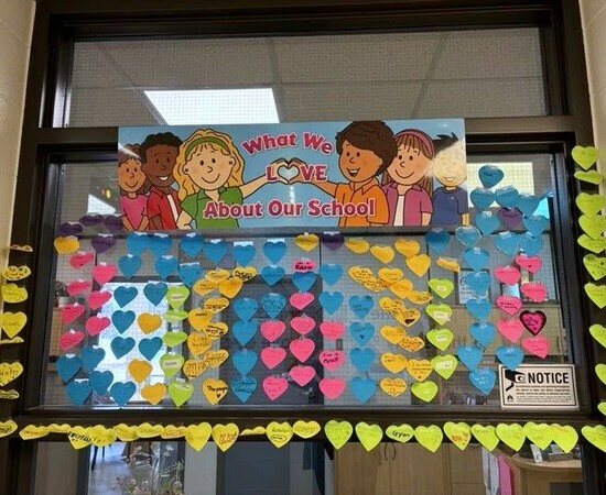 A window in a school hallway with a poster that features drawn elementary children and the text What We Love About Our School. Underneath the poster are pink, blue and yellow sticky notes with handwritten messages.