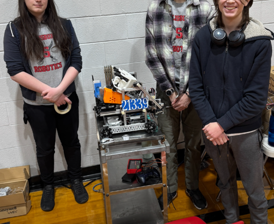 Three students stand in a gym next to a robot on a rolling cart and a red chair. The text SAL Robotics Red is in the upper left-hand corner.
