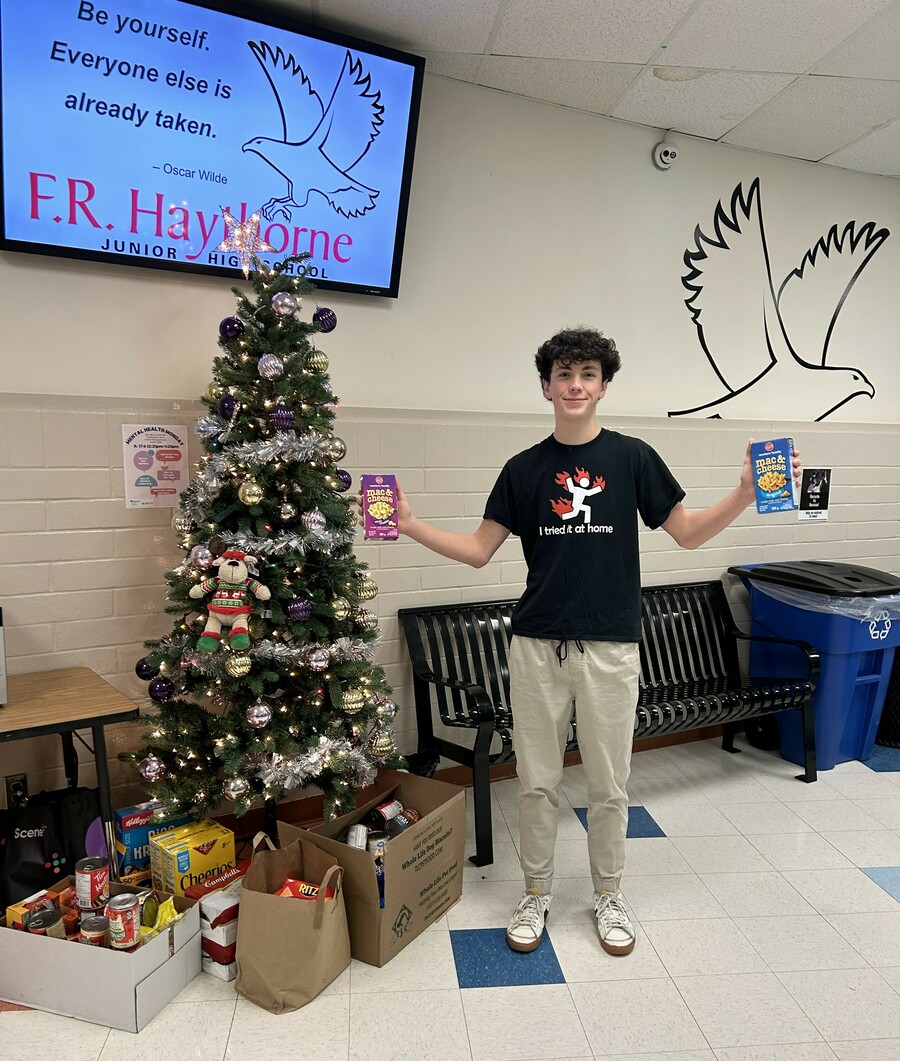A student stands in a hallway at F.R. Haythorne Junior High holding a box of macaroni in each hand. There's a decorated Christmas tree to the left with various food drive donations at the bottom of the tree, including cans of soup and cereal boxes. In the