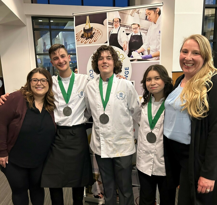 In a foyer, two school staff stand on either end of a line with three students in between them. The students are wearing white chef jackets and silver medals. Everyone smiles at the camera. In the background, there's a banner for the High School Culinary