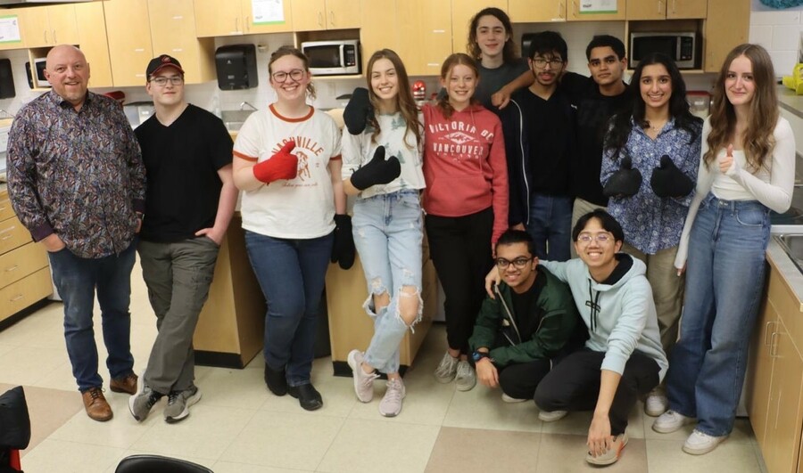 A teacher stands along with several students facing a camera in a kitchen classroom. Several students wear oven mitts and give a thumbs up.