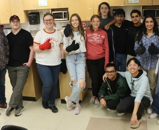 A teacher stands along with several students facing a camera in a kitchen classroom. Several students wear oven mitts and give a thumbs up.