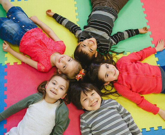 Five students lay on a multicoloured jigsaw playmat in a star shape with their heads at the centre of the image.