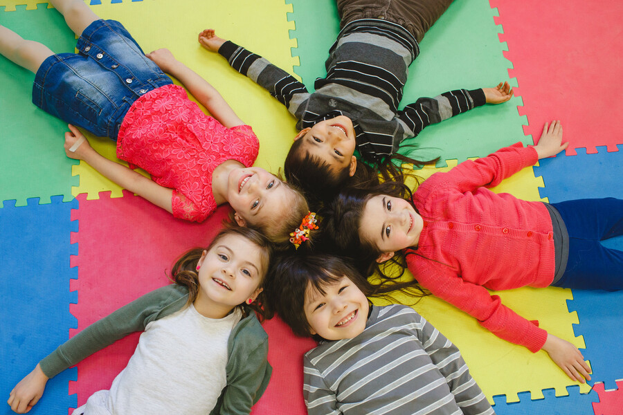 Five students lay on a multicoloured jigsaw playmat in a star shape with their heads at the centre of the image.