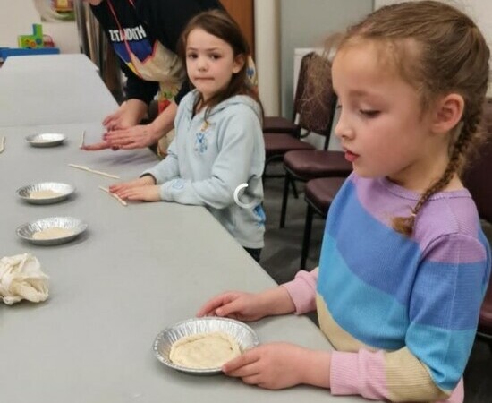 Two students sit at a long table. One student is holding a small tin of dough while the other student rolls their dough into a rope. A teacher leans over with a smile to help the student roll their dough.