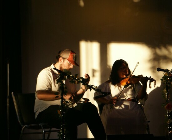 Two individuals sit in a dim room lit by a sunny window as they play fiddles together.