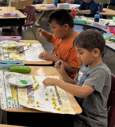 Two elementary-aged boys sit at desks creating artwork with dandelions and paint.