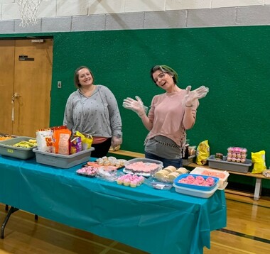 Two adults smile at the camera and stand in a school gymnasium at a table laden with chips, cupcakes and other treats.