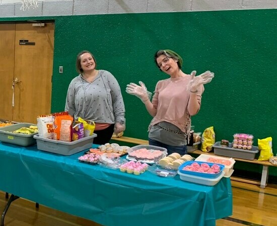 Two adults smile at the camera and stand in a school gymnasium at a table laden with chips, cupcakes and other treats.