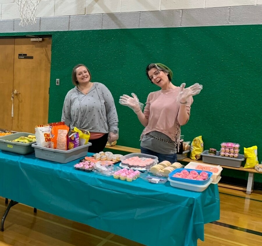 Two adults smile at the camera and stand in a school gymnasium at a table laden with chips, cupcakes and other treats.
