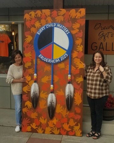 Two women stand beside a tall rectangular board painted orange with many handprints around a dreamcatcher in the centre. The text 