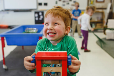 A young student wearing a green long sleeve shirt stands in a classroom holding a red toy and smiling at the camera. There's a blue sandbox elevated on table legs to their right and two other students playing to their left in the background.