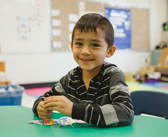 A student sits at a desk in an elementary school classroom. They smile at the camera, and they have a half-coloured piece of paper shaped like a bat under their folded arms.