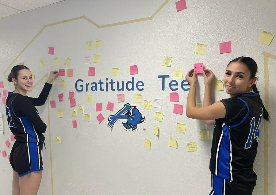 Two students stand near a wall in a hallway and post sticky notes on the wall with the text Gratitude Tee and the Ardrossan Junior Senior High logo within a taped t-shirt outline.