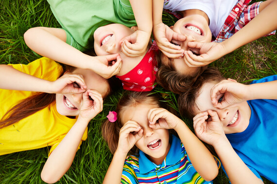 Five children in colourful clothing lay on a grassy green field looking up at the camera with smiles and their hands lifted toward their faces like binoculars.