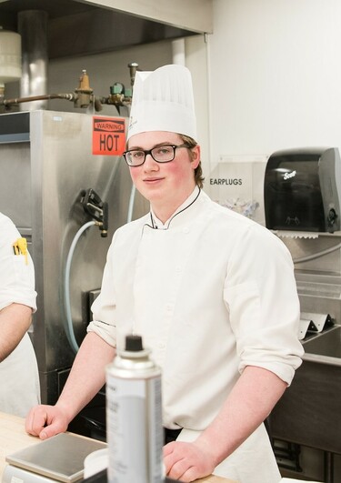 A young man wearing chef's whites and a white hat stands in an industrial kitchen