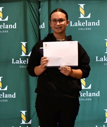 A student stands on the top step of a podium with the text Gold on its front. She smiles at the camera while holding an envelope from Skills Alberta.