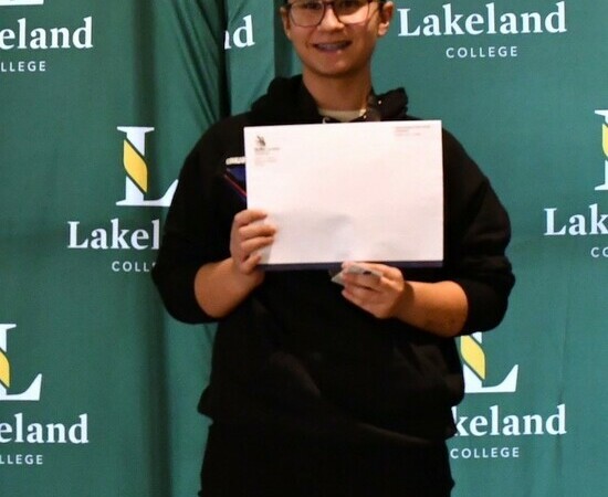 A student stands on the top step of a podium with the text Gold on its front. She smiles at the camera while holding an envelope from Skills Alberta.