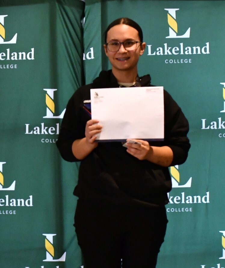 A student stands on the top step of a podium with the text Gold on its front. She smiles at the camera while holding an envelope from Skills Alberta.