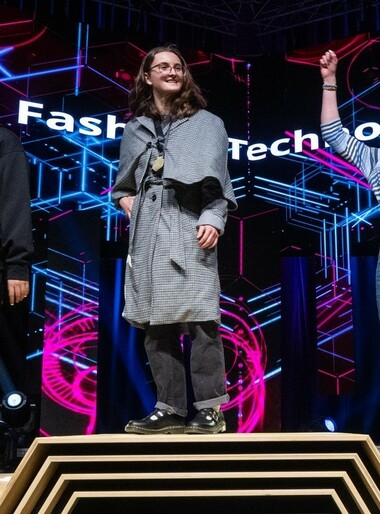 A student stands on the first-place step of a podium on a stage. They wear a gold medal and the grey plaid jacket they created.