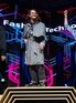 A student stands on the first-place step of a podium on a stage. They wear a gold medal and the grey plaid jacket they created.