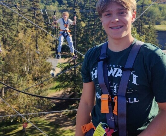In a forested area, two students wearing harnesses stand on an aerial obstacle course.