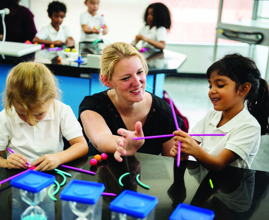 Two students take part in a STEM activity at a countertop in a classroom. A teacher crouches down next to them to help the students.