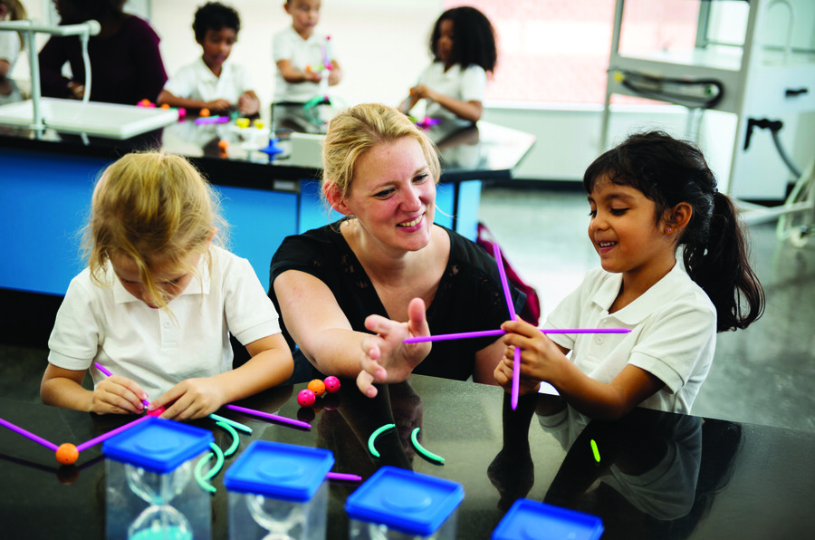 Two students take part in a STEM activity at a countertop in a classroom. A teacher crouches down next to them to help the students.