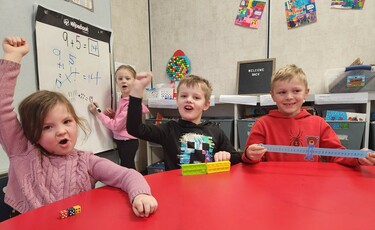 Three students sit at a red table. The student to the left holds their fist up to the air with colourful dice in front of them. The student in the middle holds their fist up in the air with two rectangular math tools in front of them. The student to the r