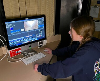 A student wearing a hoodie sits at a desk with her left hand resting on the surface and her right hand on a computer mouse. On the desk is an Apple computer monitor open to video editing software. A keyboard sits in front of the monitor.