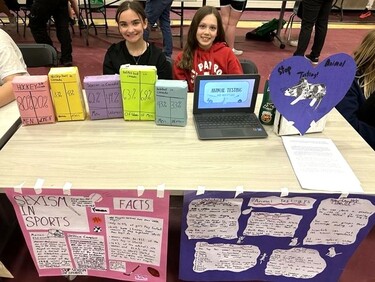 Two students sit at a table with their human rights projects in front of them. The student on the left explored sexism in sports and the students on the right explored animal testing.