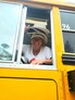 A woman wearing multiple hats and a white T-shirt smiling and sitting behind the steering wheel of a yellow school busing looking out the open side window.