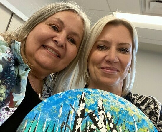 Two women stand together behind a painting of leaf-less birth trees against a blue and green sky