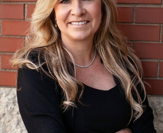 Headshot of a woman with long blonde hair wearing a dark top standing in front of a brick wall
