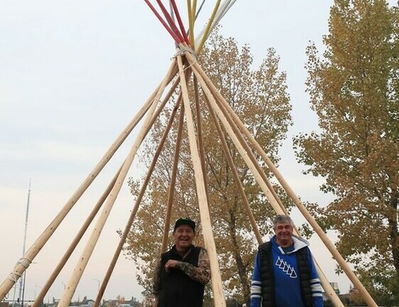 Two men stand outside beside a tipi, poles only