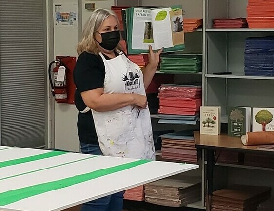 Woman wearing a face mask and apron holds up a book in front of a class
